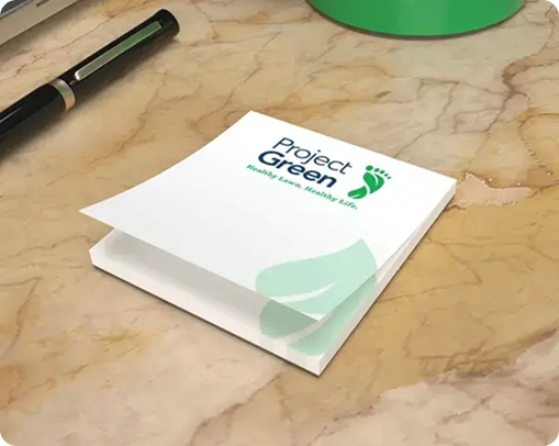 A stack of white sticky notes with Project Green and a green footprint logo on top, placed on a marble desk next to a black pen—perfect as branded products for eco-conscious promotional campaigns.
