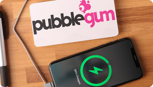 A smartphone charging on a wooden surface displays a 95% charged screen. Next to it is a card with the pubblegum logo in black and pink text, alongside custom merchandise and a pen partially visible on the left side.