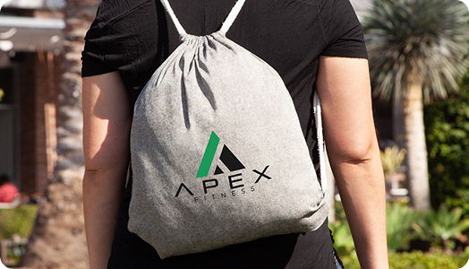 Person wearing a light gray drawstring backpack with the Apex Fitness logo—one of our premium promotional products—standing outdoors near palm trees and greenery.