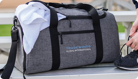 A gray duffel bag with black straps and TRADEWINDS GLOBAL INTERNATIONAL printed on the side—an example of branded products—sits on a bench. A white towel is draped over it and someone ties their black athletic shoe nearby.