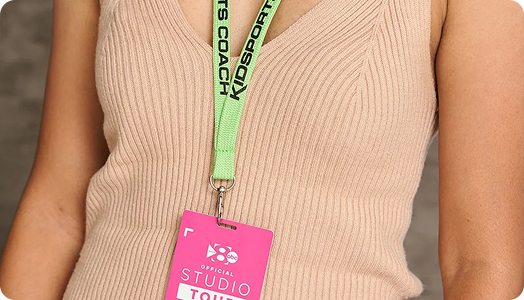 A person wearing a beige ribbed sleeveless top and a green lanyard that reads SPORTS COACH, holding a pink badge labeled STUDIO TOUR—showcasing custom merchandise as eye-catching promotional products.