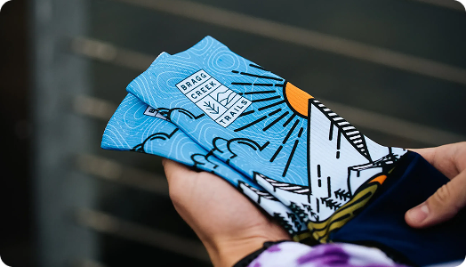 A person holds colorful socks featuring a blue and white mountain design with a sun, clouds, and the text BRAGG CREEK TRAILS—perfect custom merchandise for trail lovers.