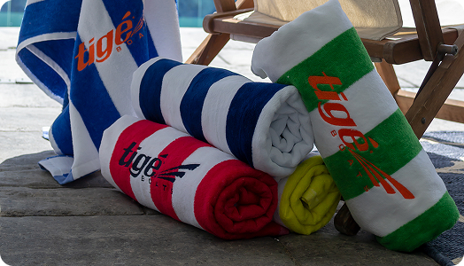 Four rolled beach towels—blue, red, yellow, and green—each featuring the “Tigé Boats” logo as stylish branded products, are placed on a stone surface next to a wooden chair. One blue and white towel is draped over the back of the chair.