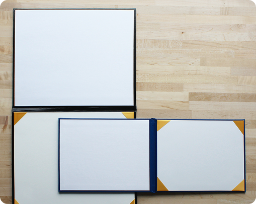 Three empty photo albums with blank white pages and gold photo corners—perfect as branded products or custom merchandise—are arranged on a light wooden surface, with two albums open and one closed.