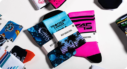 A variety of colorful socks with different patterns and designs are laid out on a white surface. Some socks, labeled BEST SOCKS EVER, showcase custom merchandise with tropical scenes, sports themes, and bold graphics.