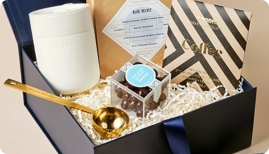 A gift box featuring custom merchandise: a white travel mug, gold coffee scoop, Blue Velvet coffee, chocolate-covered espresso beans, and a patterned chocolate bar—all nestled in shredded paper for a premium promotional products experience.