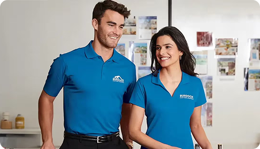 Two people in matching blue polo shirts featuring branded products stand side by side indoors, smiling and looking to the left. A white wall behind them displays various photos and notes.