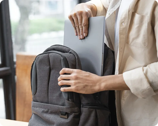 A person in a light-colored shirt is placing a silver laptop and other custom merchandise into the main compartment of a gray backpack indoors near a window.