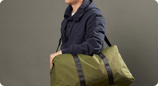 A person wearing a dark blue hoodie carries an olive green duffel bag with black straps—an ideal choice for custom merchandise or branded products—over their shoulder against a plain gray background.