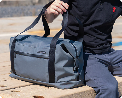 A person in a black shirt and blue pants sits on a wooden surface, holding the handles of a large gray duffel bag with black straps—a stylish piece of custom merchandise with a front zipper pocket.