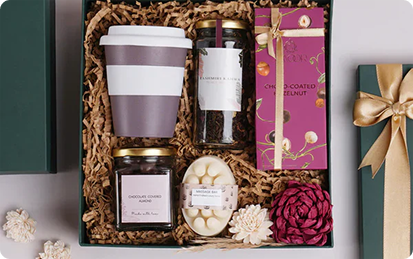 A gift box containing branded products: a reusable cup, two labeled glass jars, a pink box of chocolate-coated hazelnuts, a round soap, and decorative flowers, all arranged on shredded paper.