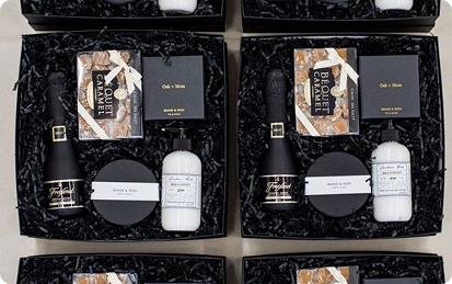 Four black gift boxes filled with caramel candy packs, candles, white lotion bottles, and small black custom merchandise, all nestled in black shredded paper.