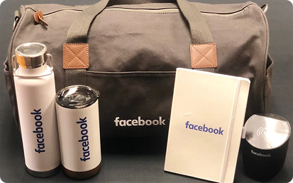 A gray duffel bag, water bottle, tumbler, notebook, and wireless charger—premium promotional products branded with the Facebook logo—are displayed on a sleek black surface.