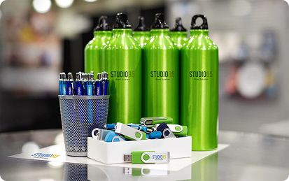A row of green water bottles, a metal cup holding blue pens, and a box of assorted USB flash drives are displayed on a table. These branded products, labeled STUDIO85, make perfect promotional products for your business.