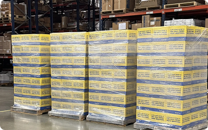Stacks of pallets wrapped in plastic, each loaded with boxes labeled “TRUST HOSPITAL” in yellow and blue, showcase branded products arranged in a large warehouse with shelves and other packages in the background.