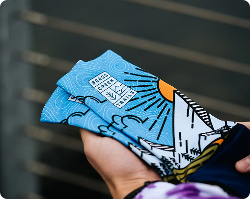A hand holds up a pair of blue socks decorated with a mountain, sun, and tree design, featuring the words BRAGG CREEK TRAILS. These custom merchandise socks are shown with a blurred railing in the background.