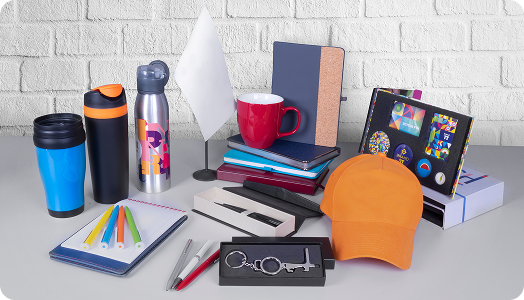 A selection of colorful office supplies and custom merchandise, including notebooks, pens, mugs, water bottles, an orange cap, keychains, colored pencils, a blank flag, and a box of buttons displayed on a desk.