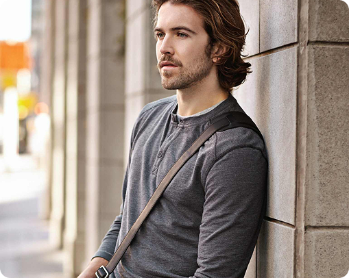 A man with wavy brown hair and a mustache leans against a stone wall outdoors, wearing a gray long-sleeve henley shirt and a crossbody bag—perfect for carrying custom merchandise or other promotional products—looking thoughtfully into the distance.